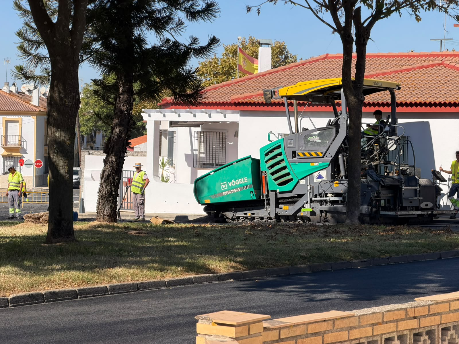 Una extendedora de asfalto verde y amarilla junto a operarios con chalecos reflectantes en una calle con árboles y casas de tejado rojo.