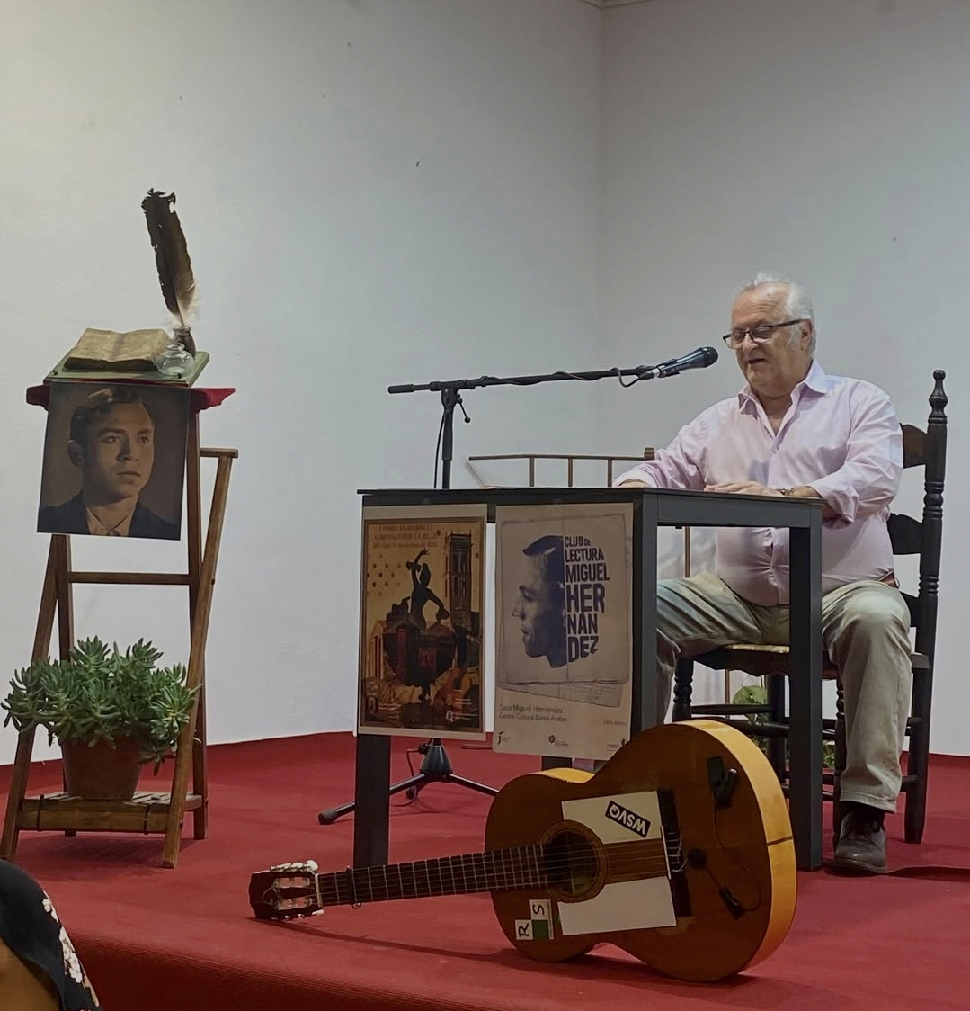 Un hombre mayor sentado tras una mesa con micrófono en un escenario con carteles, una guitarra y un atril con un retrato.