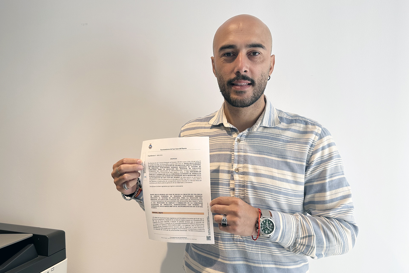 Hombre calvo con barba sostiene un documento del Ayuntamiento de San Juan del Puerto frente a una pared blanca.