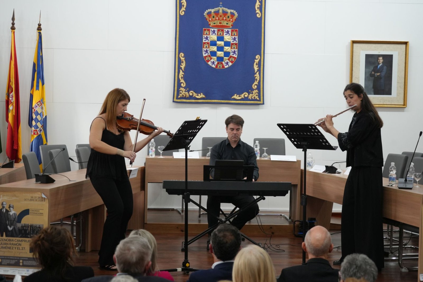 Dos músicas con violín y flauta y un teclista tocan ante un público en una sala con banderas y un escudo.