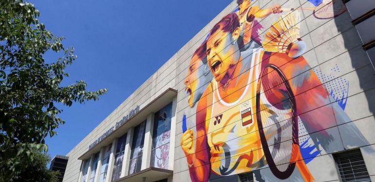 Fachada de un edificio deportivo con un gran mural de una jugadora de bádminton gritando, raqueta en mano, bajo un cielo azul.
