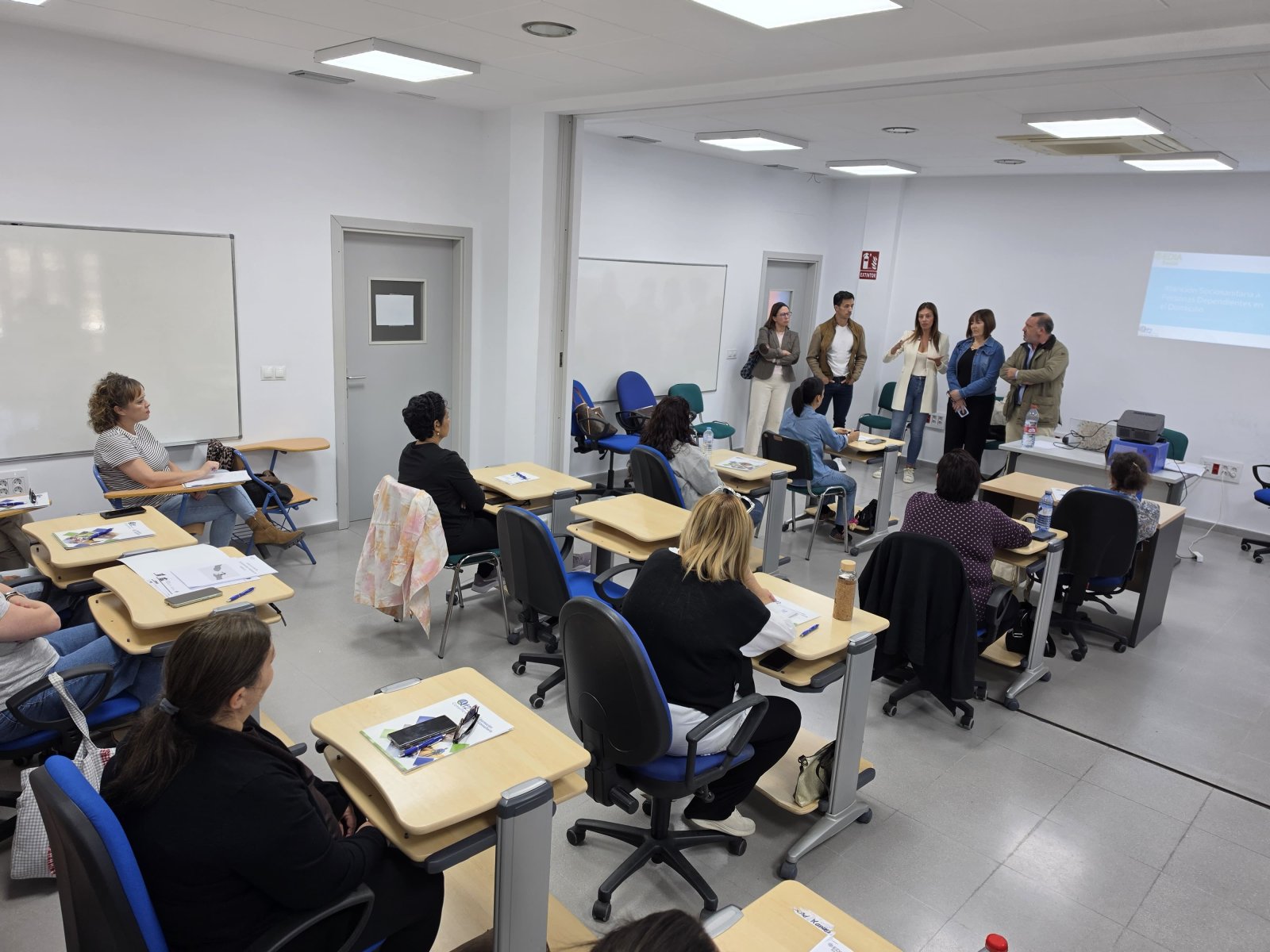 Personas sentadas en pupitres de un aula miran a un grupo de pie junto a una pantalla de proyección.