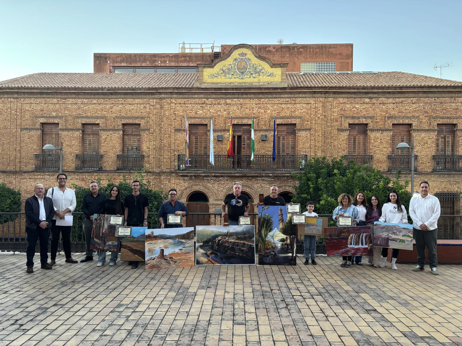 Un grupo de personas posa en una plaza con cuadros y diplomas ante un edificio de ladrillo con banderas.