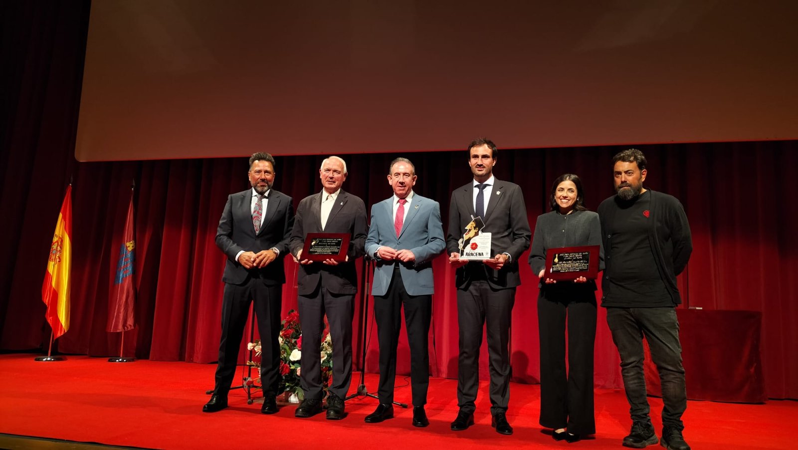 Seis personas posan en un escenario con fondo rojo, banderas de España y Andalucía, y algunas sostienen placas y un trofeo.