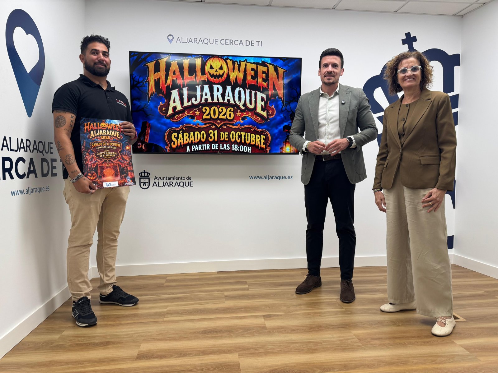 Tres personas posan de pie en una sala con una pantalla que anuncia Halloween en Aljaraque y uno de ellos sostiene un cartel.