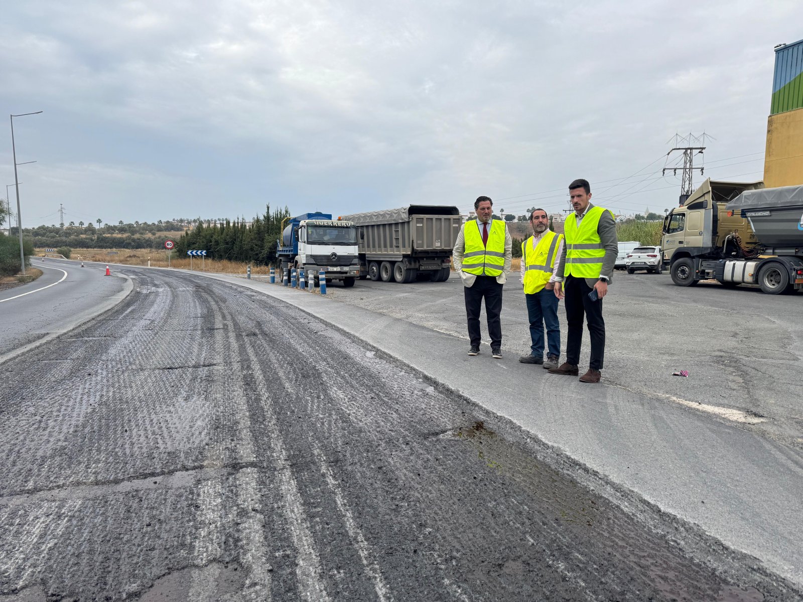 Tres hombres con chalecos reflectantes de pie junto a una carretera en obras, con camiones y una señal de 40 al fondo.