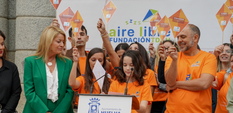 Una niña lee un discurso en un atril ante varias personas con camisetas naranjas y carteles de colores.