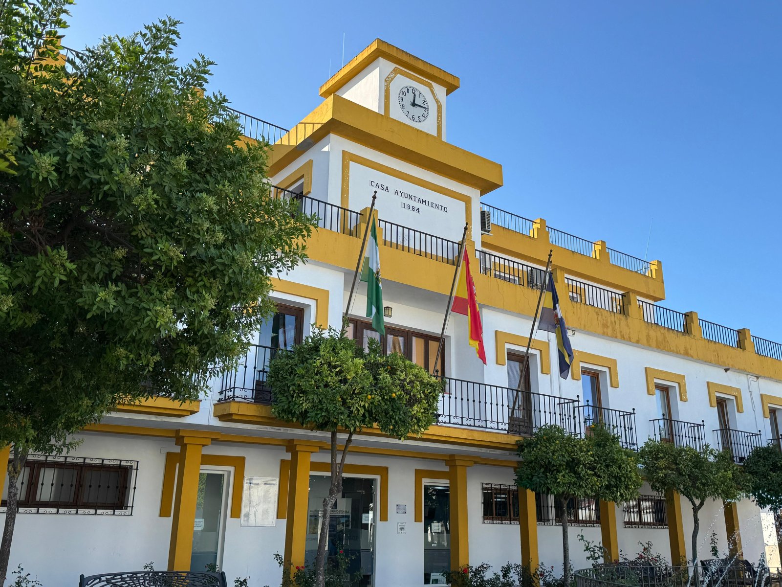 Edificio blanco con remates amarillos, torre con reloj y la inscripción Casa Ayuntamiento 1984, con tres banderas y árboles delante.