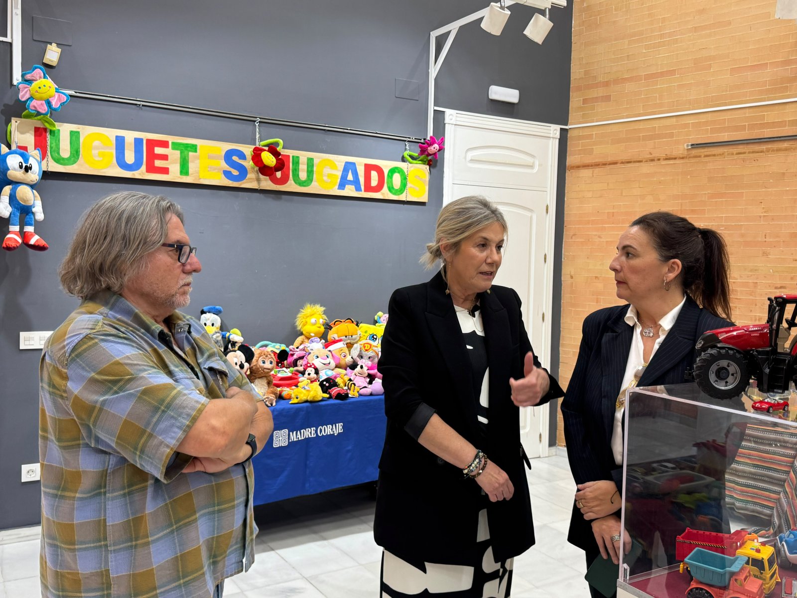 Tres personas conversan ante una mesa con peluches y un cartel de letras de colores que dice JUGUETES JUGADOS.