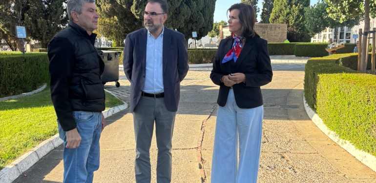 Tres personas de pie conversando en un camino de tierra entre setos y árboles, junto a una papelera.