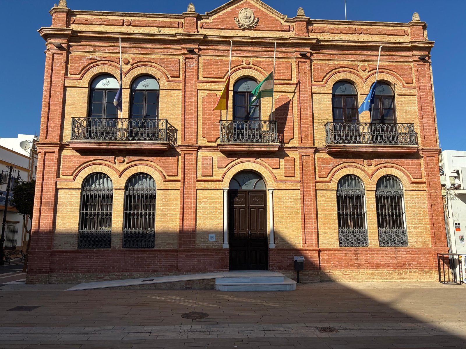 Edificio de ladrillo rojo con arcos, balcones y banderas en la fachada, frente a una plaza pavimentada.