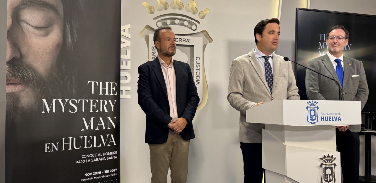 Tres hombres trajeados posan junto a un cartel de la exposición The Mystery Man y un atril con el escudo de Huelva.