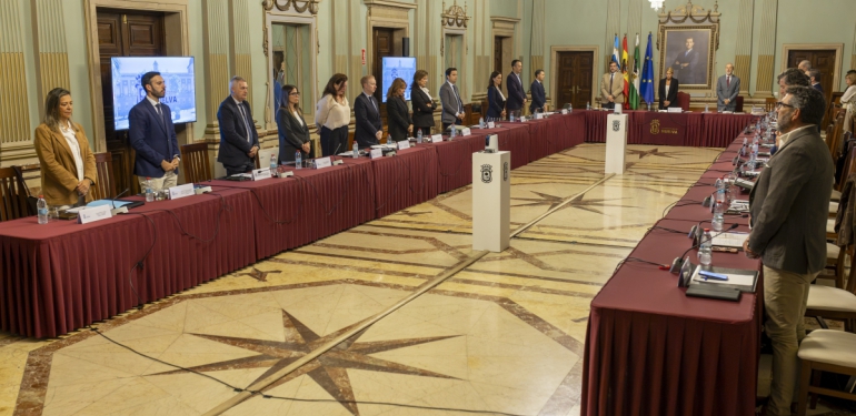 Personas de pie junto a una mesa alargada con mantel granate en una sala amplia con suelo de estrellas y banderas al fondo.