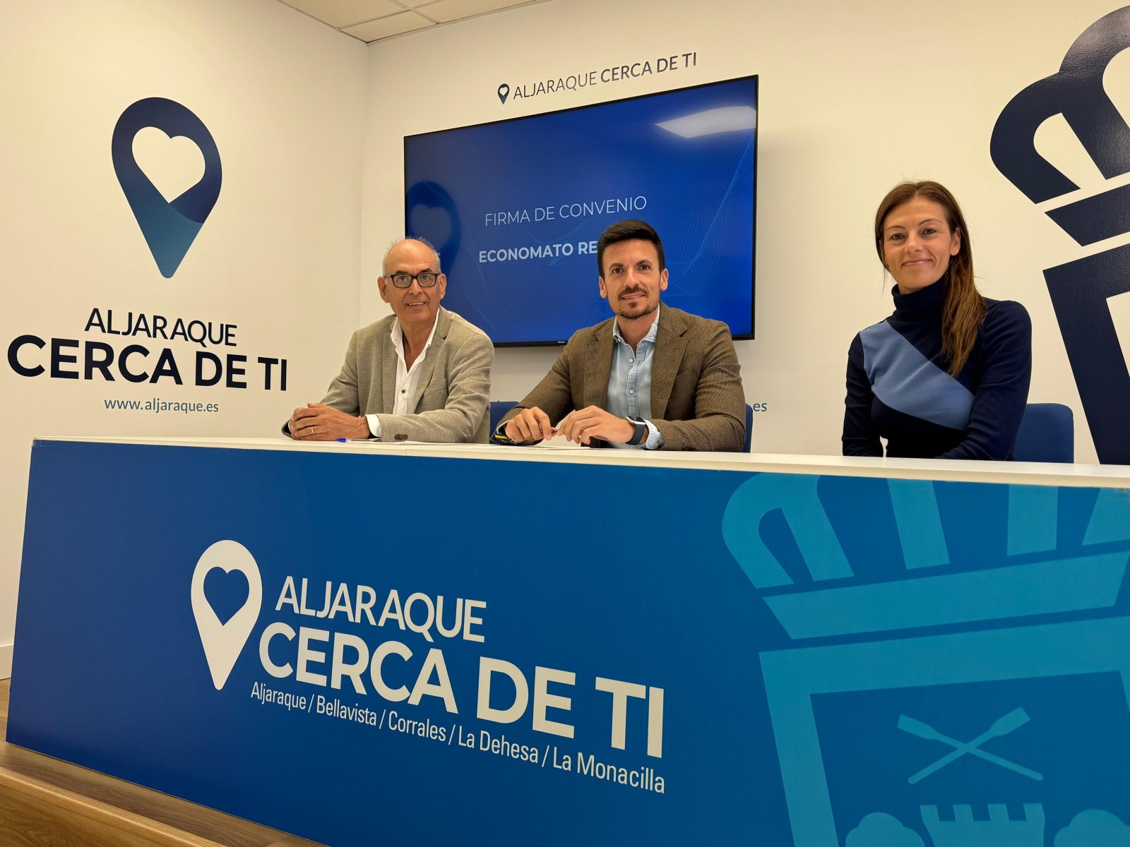 Tres personas sentadas tras una mesa azul con el lema Aljaraque Cerca de Ti y una pantalla que dice Firma de Convenio.
